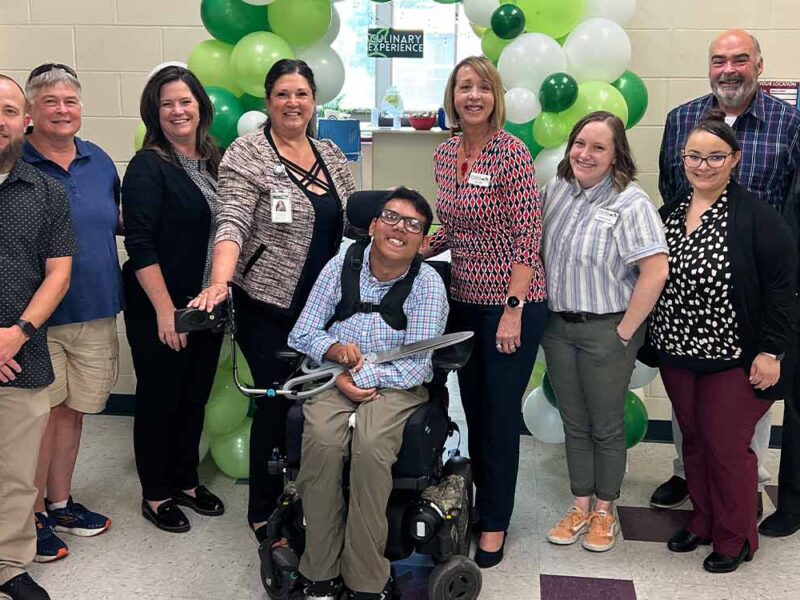 Student in wheelchair hold scissors with other people standing to sides at ribbon cutting ceremony